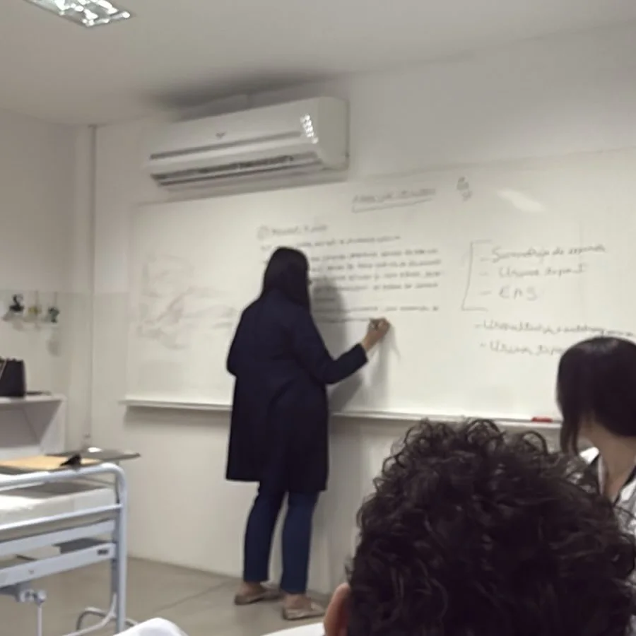 A Dra. Patricia Rennée, de costas, escreve no quadro branco durante uma aula; alunos de jaleco assistem.