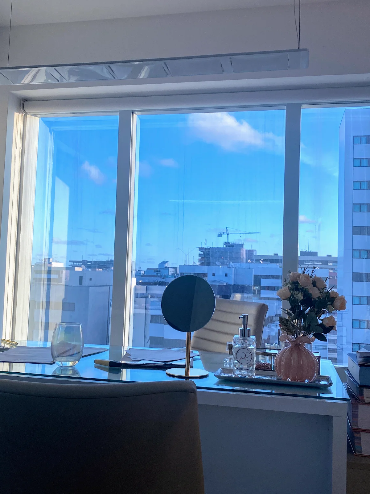 Mesa do consultório diante de uma janela ampla, com os prédios de Jatiúca e o céu azul ao fundo.