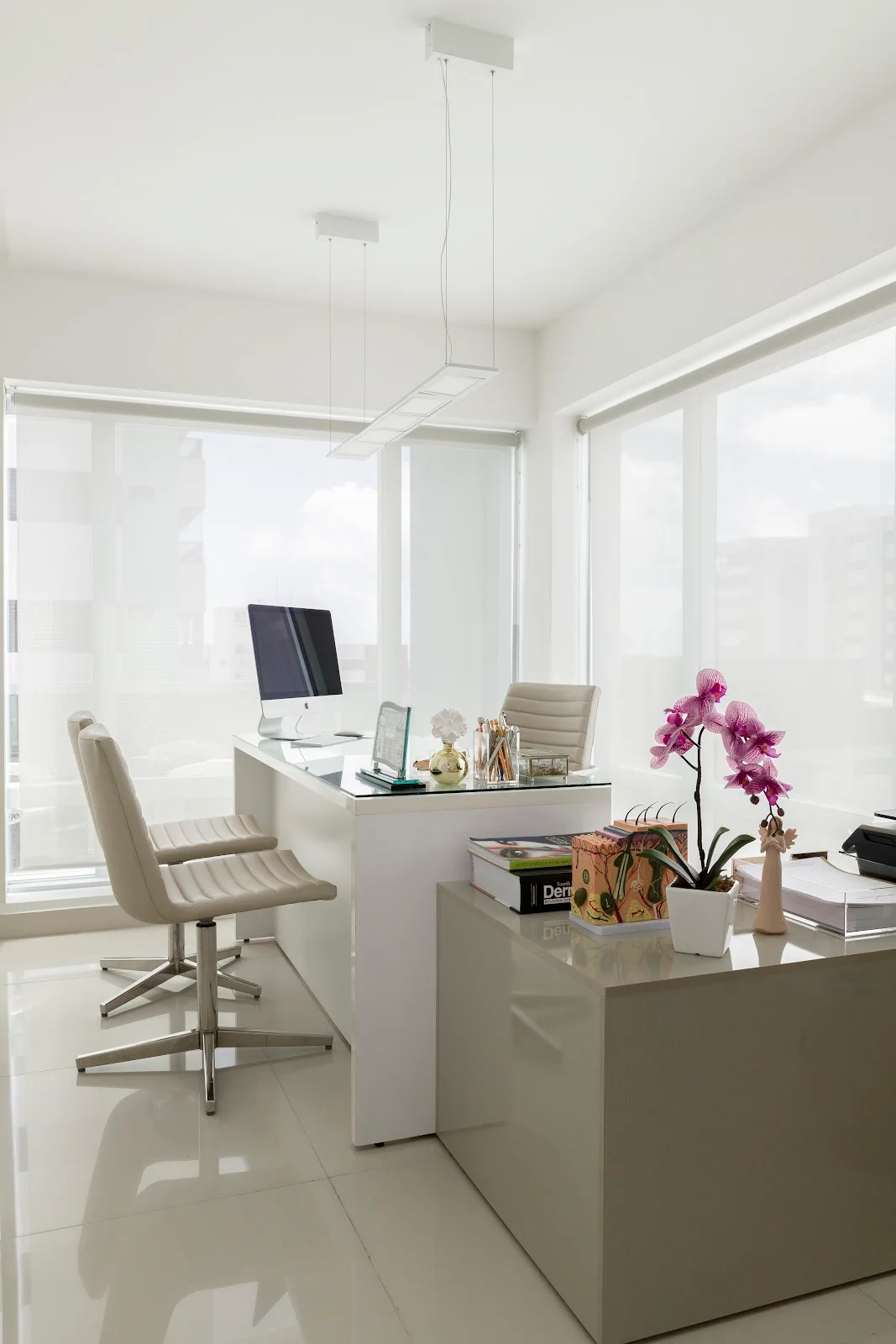 Consultório claro com mesa branca, duas cadeiras para o paciente e uma orquídea rosa.