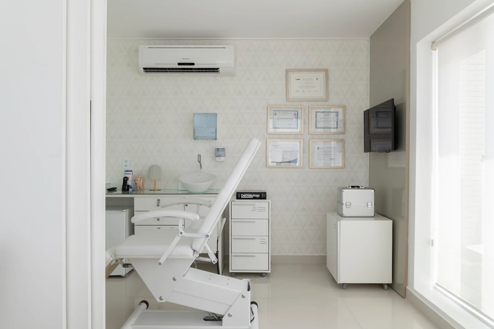 Sala de procedimentos com maca branca, armário de gavetas e certificados na parede.